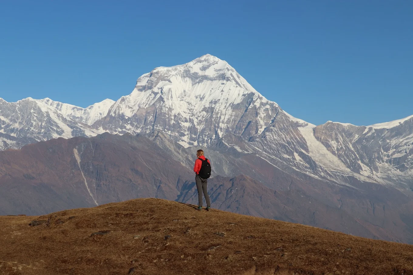 A trekker on a dry ridge looking toward a snow-covered peak